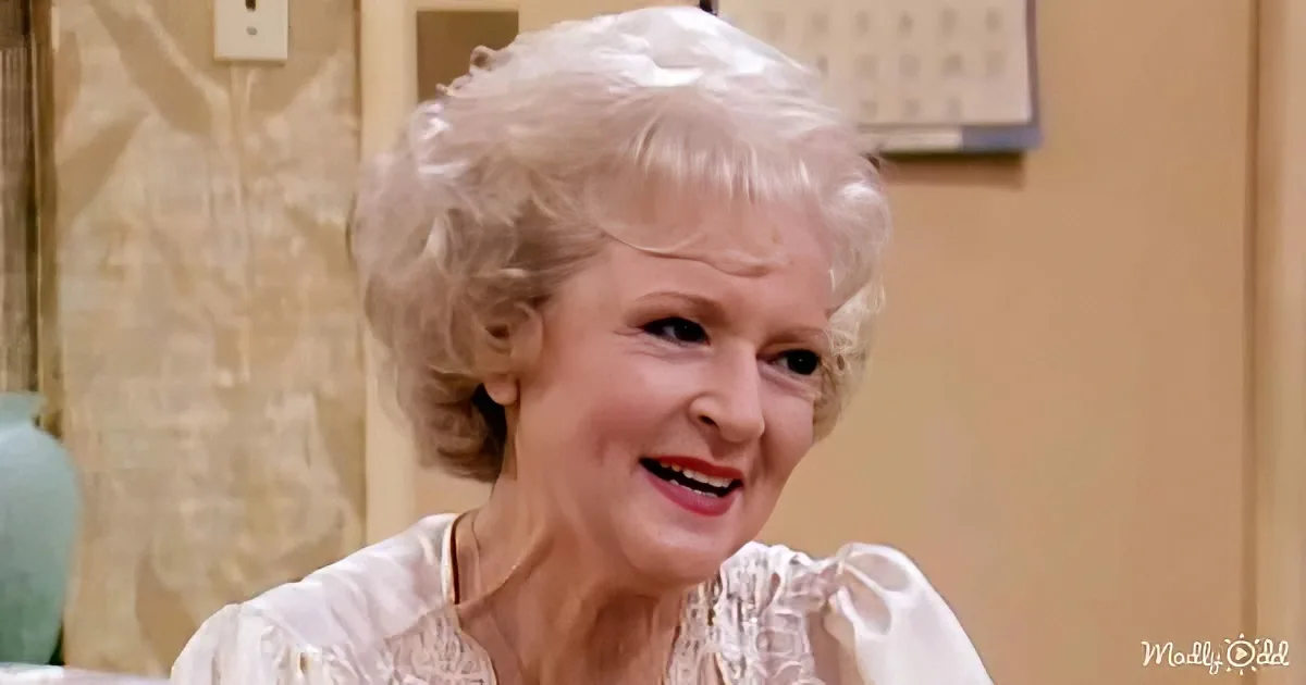 An older woman with short, curly blonde hair smiles warmly, wearing a white blouse with ruffled sleeves.