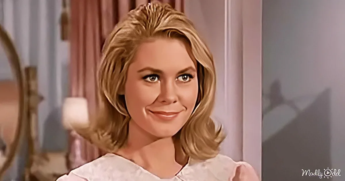A woman with blonde hair in a soft bob, wearing a white top, smiles warmly in a softly lit room.