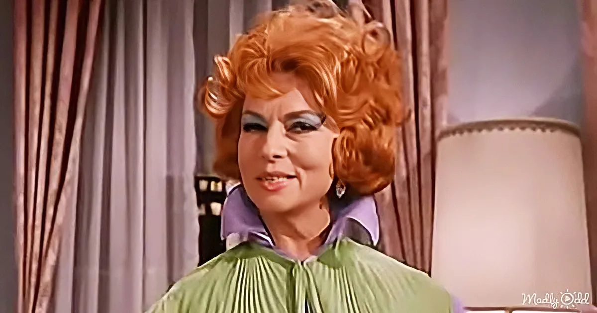 A woman with red curls and green pleated dress smiles in a room with curtains.