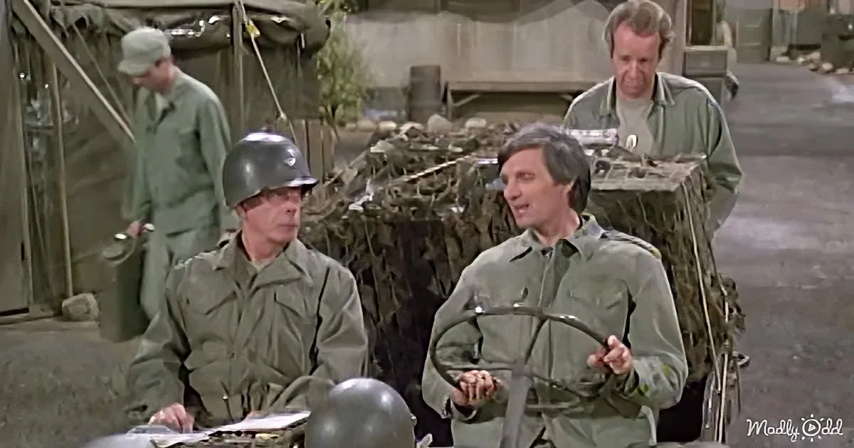 A man in a military uniform holds a curved object while another in a helmet sits at a desk, with others in military...