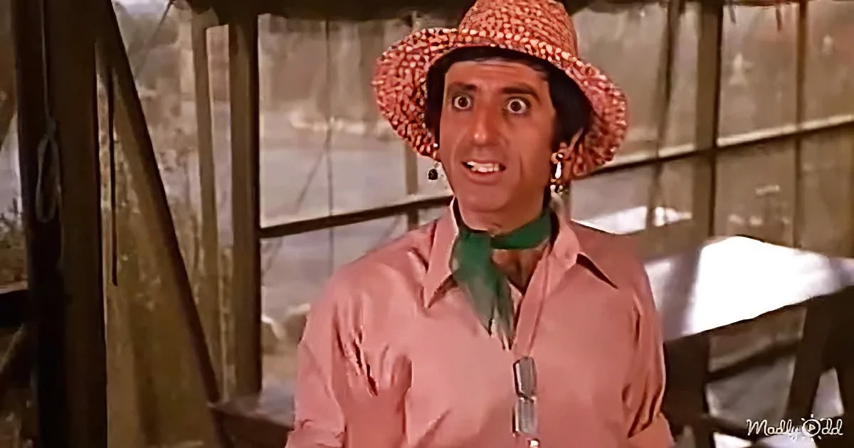 A man in a pink shirt and red hat with a surprised expression stands in a rustic setting.