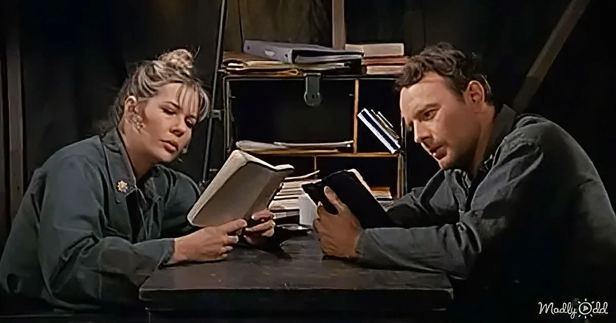 A man and woman in green uniforms sit at a desk, each holding a book, seemingly engaged in a discussion.