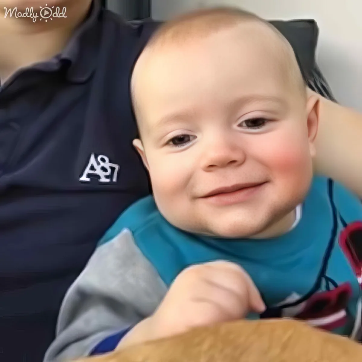 A smiling baby in a blue bib, wearing a gray and blue outfit, sits on someone's lap.