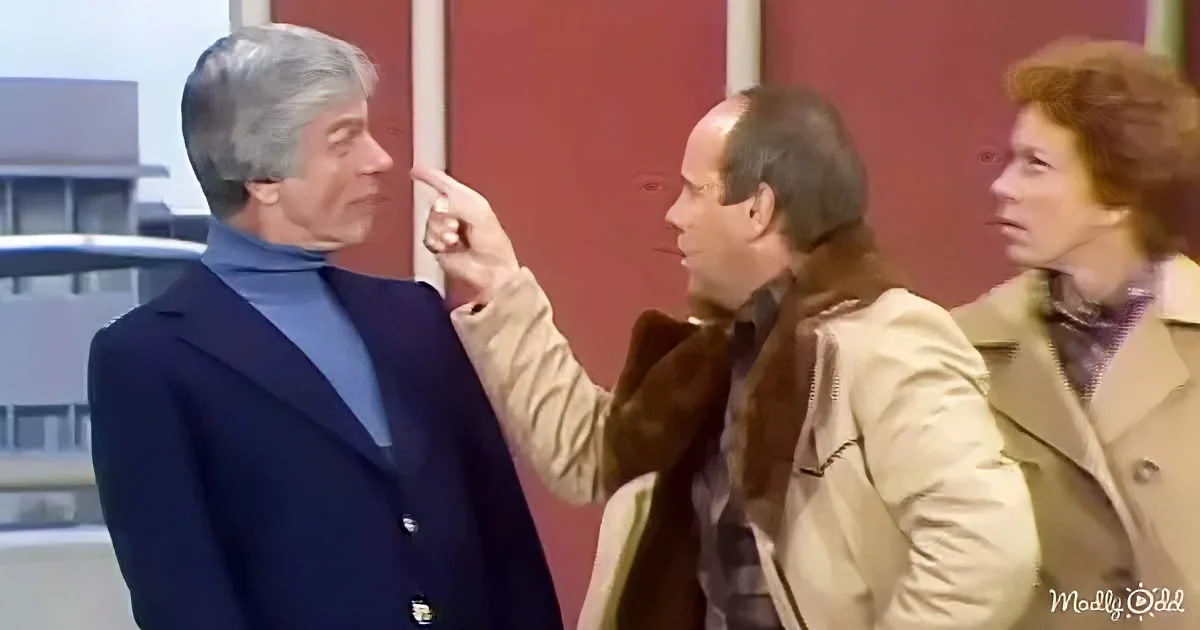 A man in a blue suit is being pointed at by another man in a beige coat, with a woman in a beige coat standing nearby.