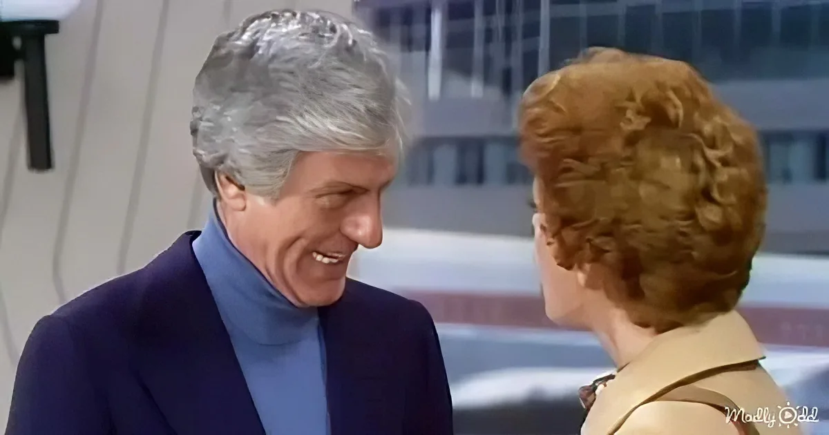 A man in a blue turtleneck and dark jacket smiles at a woman with curly red hair in a beige jacket.