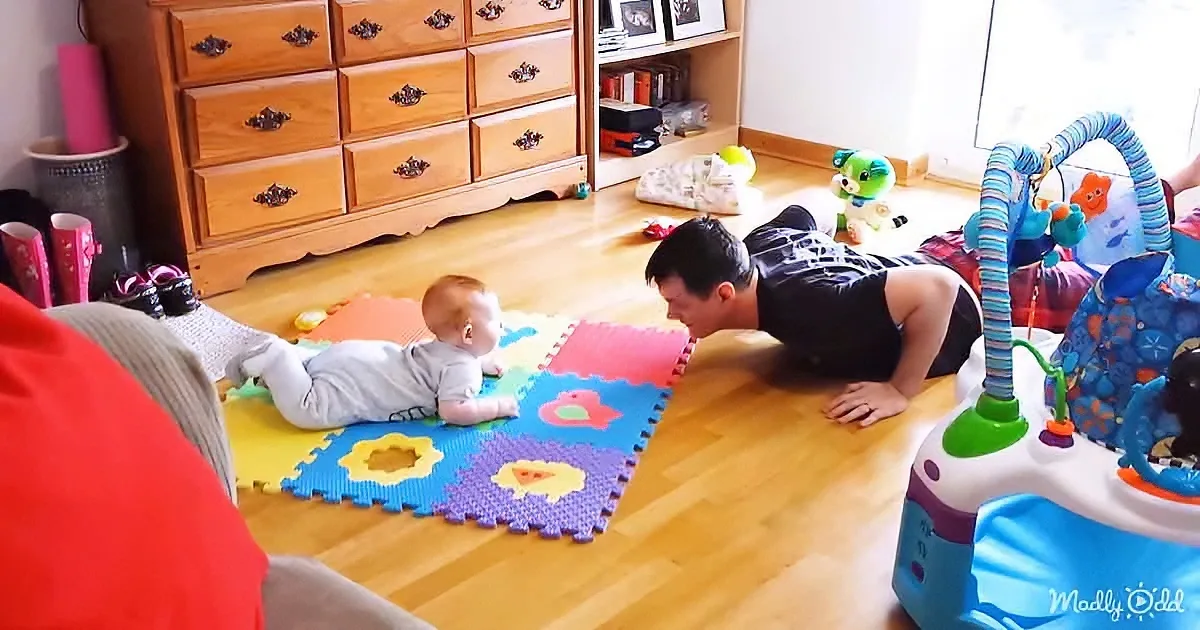 A baby lies on a colorful mat while a man does push-ups nearby, both on a wooden floor.