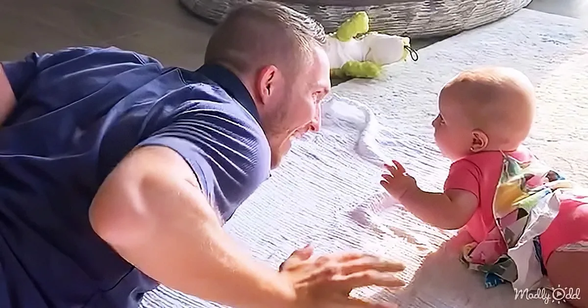 A man in a blue shirt is lying on the floor, smiling at a baby in a pink outfit who is reaching out towards him.