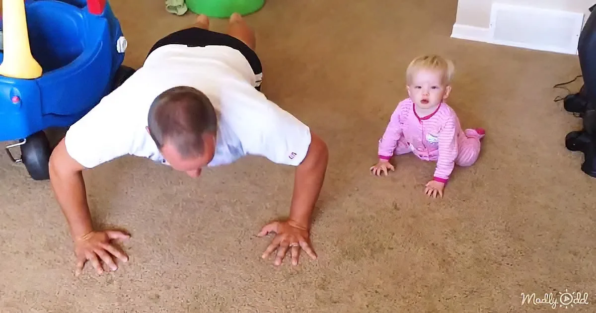 A man does push-ups on the floor while a baby in pink pajamas watches.