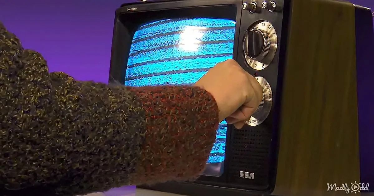 A hand in a knitted sweater adjusts the dial on a vintage television with static on the screen.