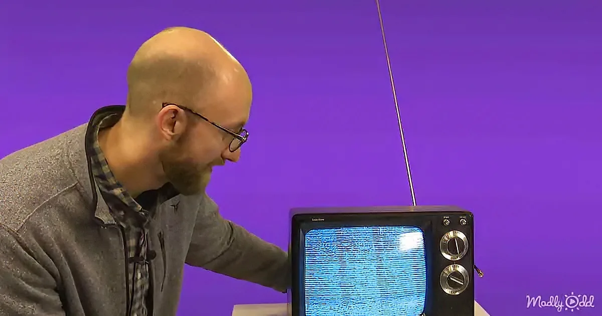 A man in glasses leans over a vintage TV with static, against a purple background.