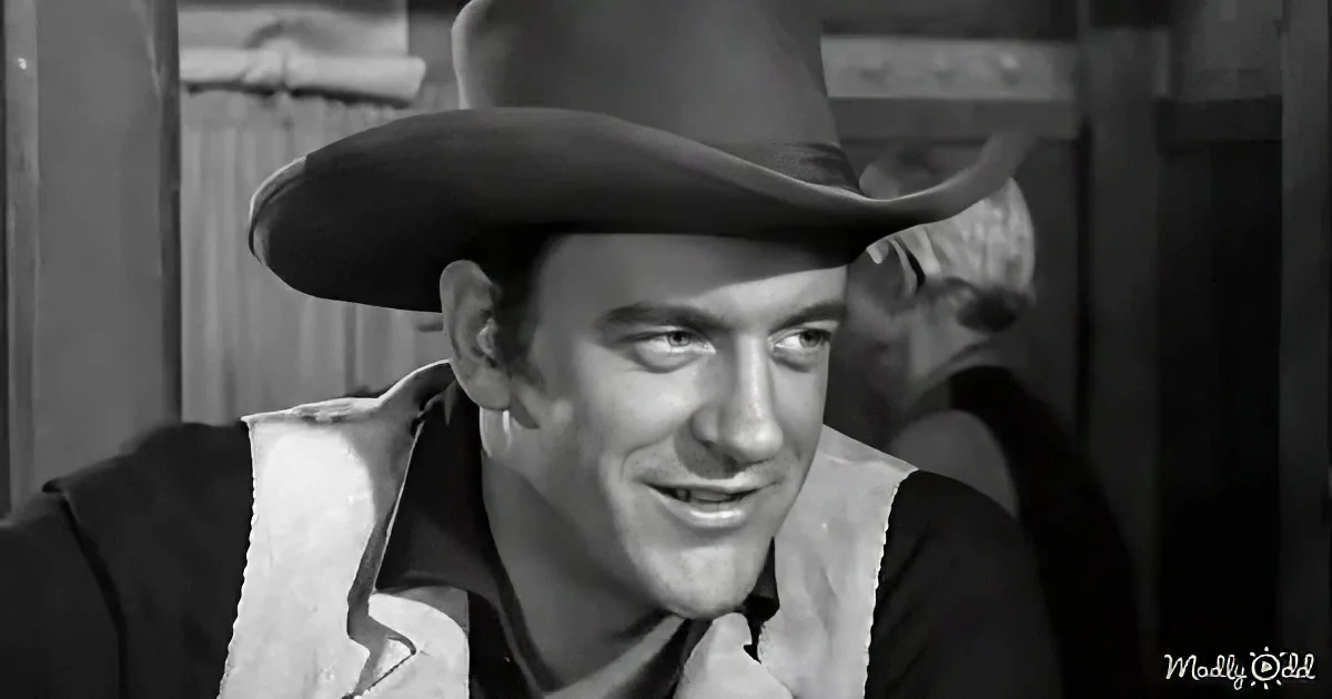 A man in a cowboy hat and vest smiles, with another person in the background.