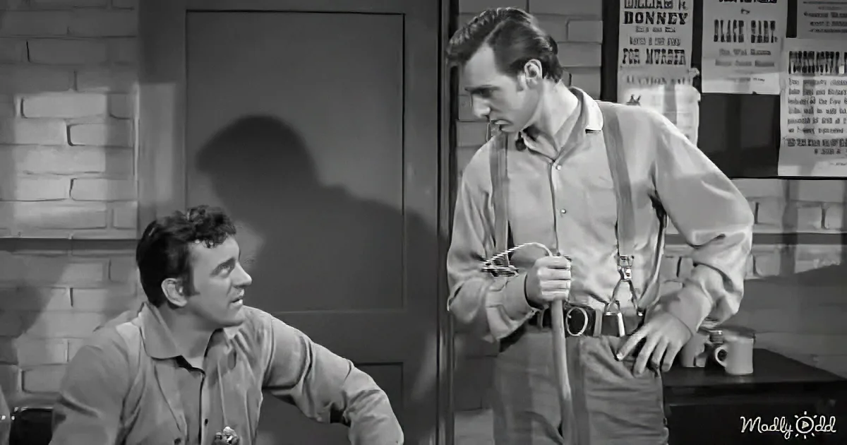 A man in suspenders holds a rope, standing next to another man in a dimly lit room.