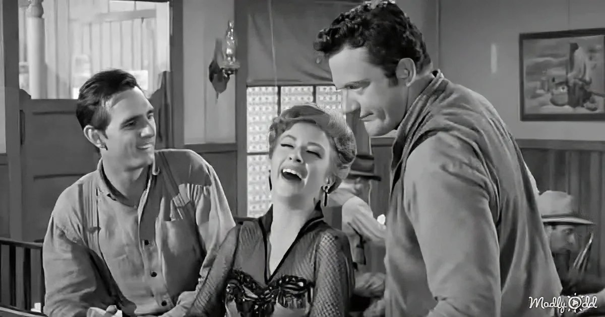 A woman laughs joyfully, surrounded by two men in a rustic setting, possibly a bar or saloon.