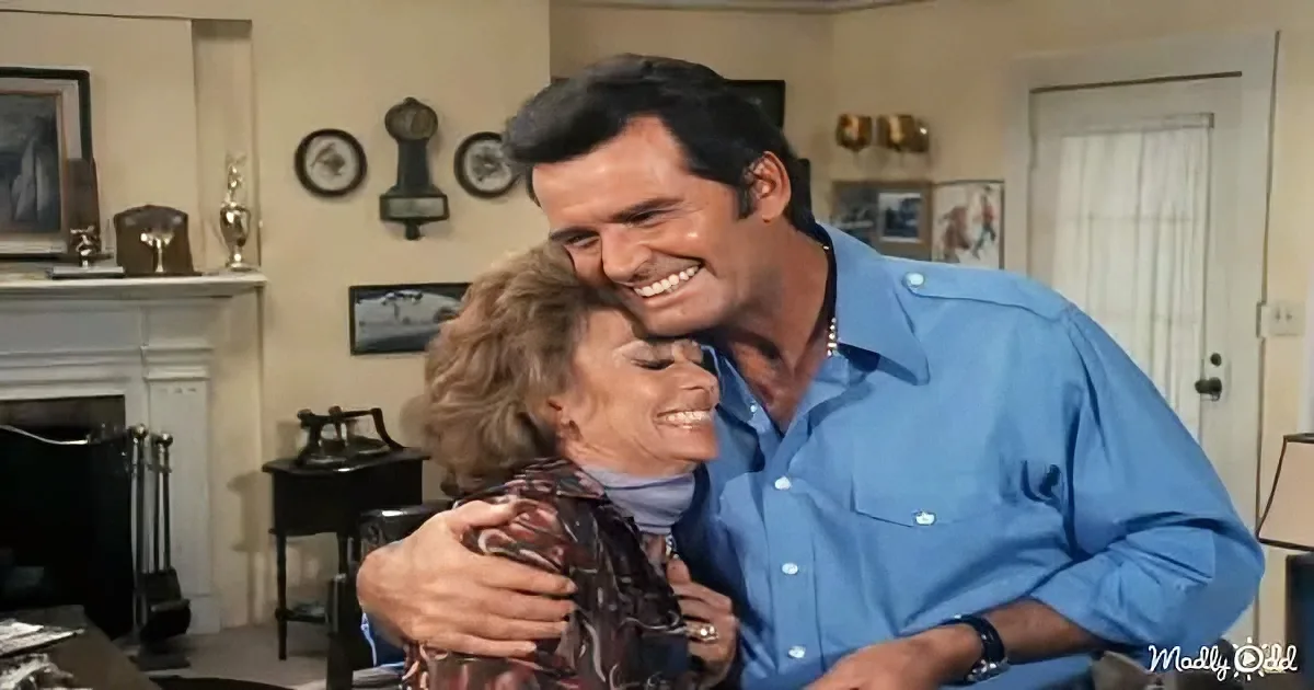 A man in a blue shirt hugs a woman in a patterned shirt, both smiling warmly in a cozy living room.
