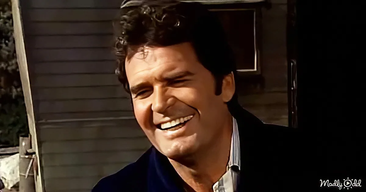 A man with dark, curly hair smiles broadly, wearing a dark jacket and a light shirt, standing outdoors near a building.