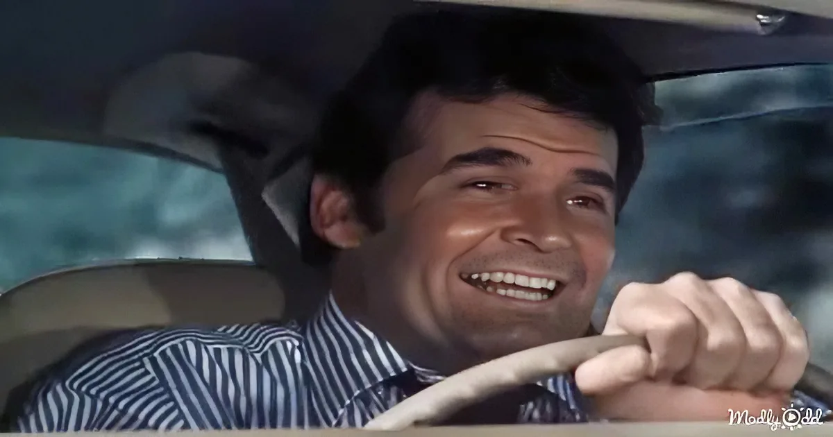 A man in a striped shirt drives a car, smiling with his hands on the steering wheel.