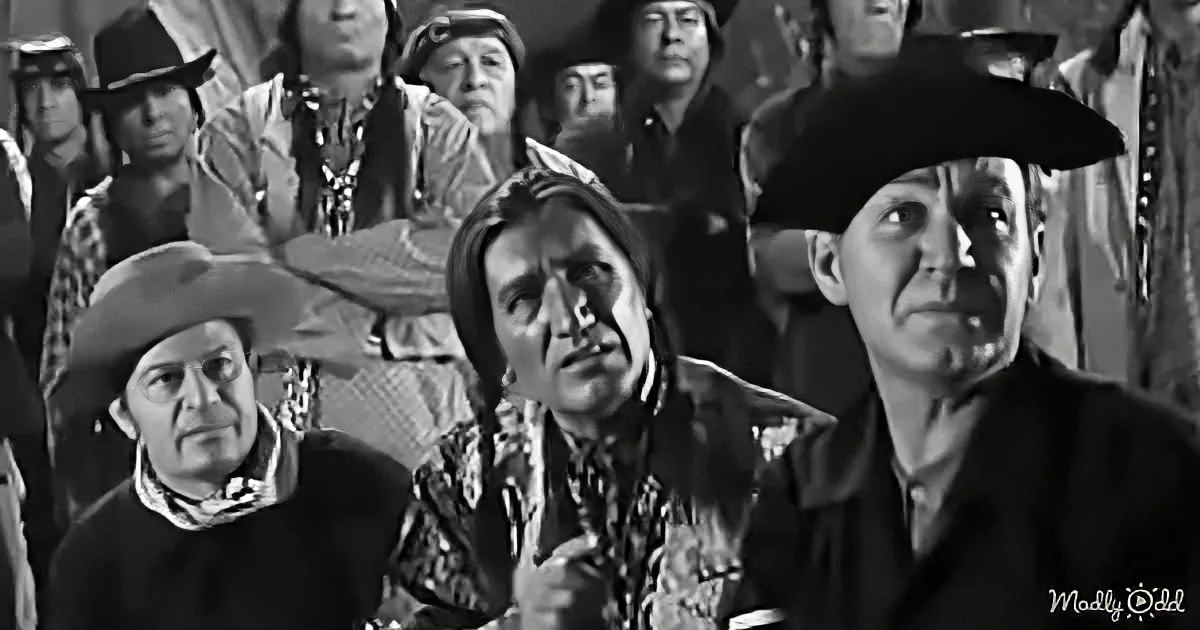 A man in a cowboy hat and patterned shirt stands among a crowd of Native Americans in a tense scene.
