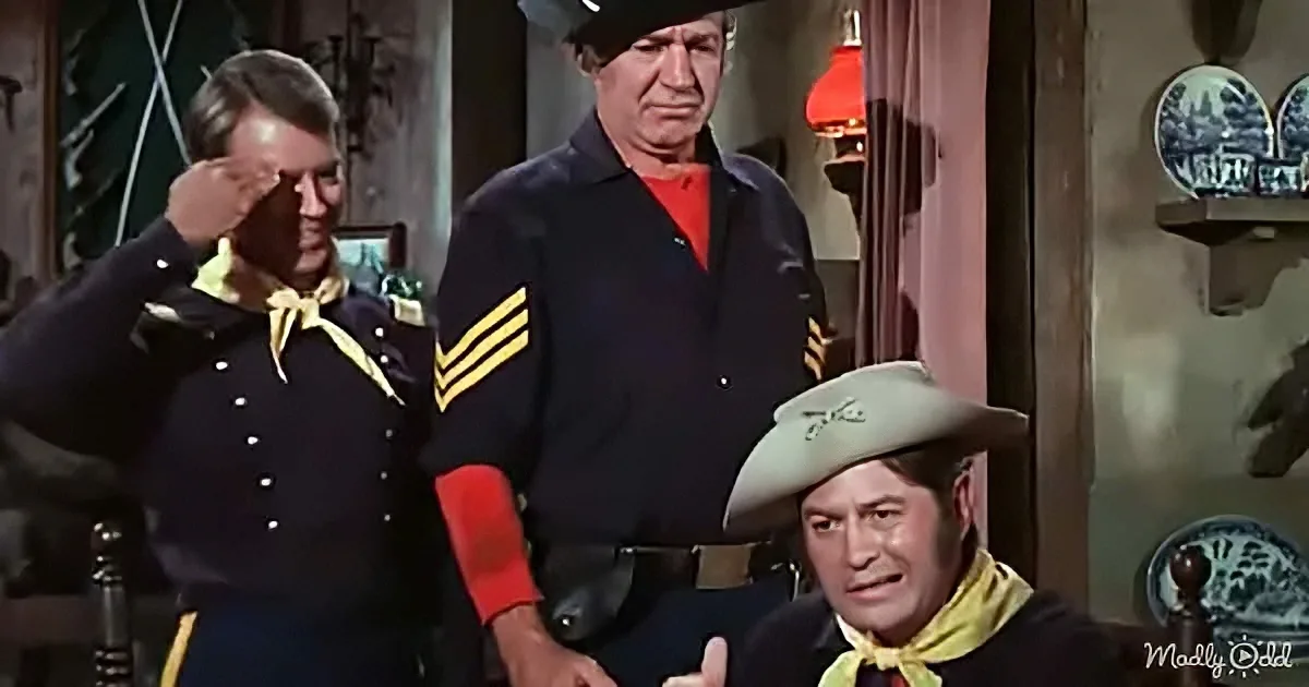 A man in a cowboy hat salutes while another in a uniform stands beside him, with a third man seated in the foreground.