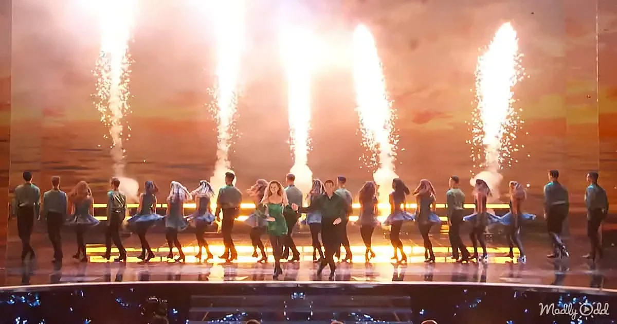 A group of dancers in green outfits perform on stage with bright, fiery fountains in the background.