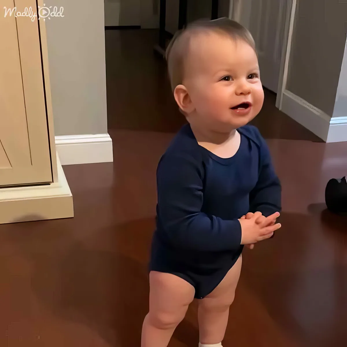 A baby in a blue onesie stands on a hardwood floor, hands clasped, looking to the side with a curious expression.