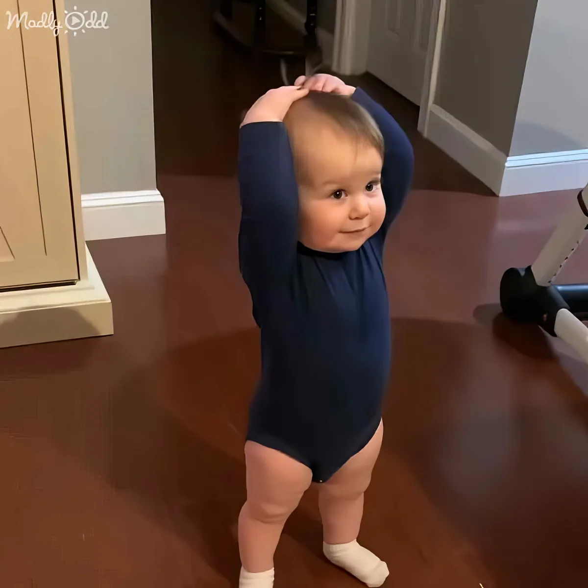 A baby in a dark blue onesie stands on a wooden floor, holding its head with both hands.
