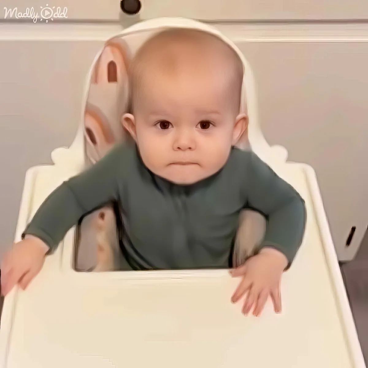 A baby in a green shirt sits in a high chair, looking forward with hands on the tray.