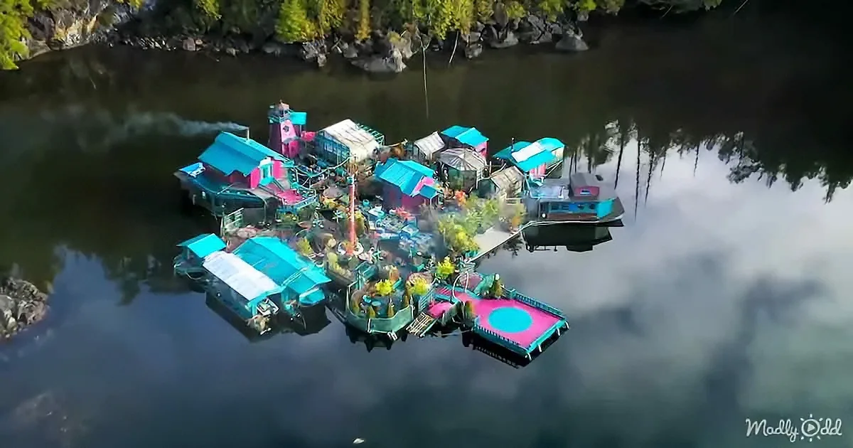 A colorful floating village with various buildings and a trampoline on a calm river surrounded by trees.