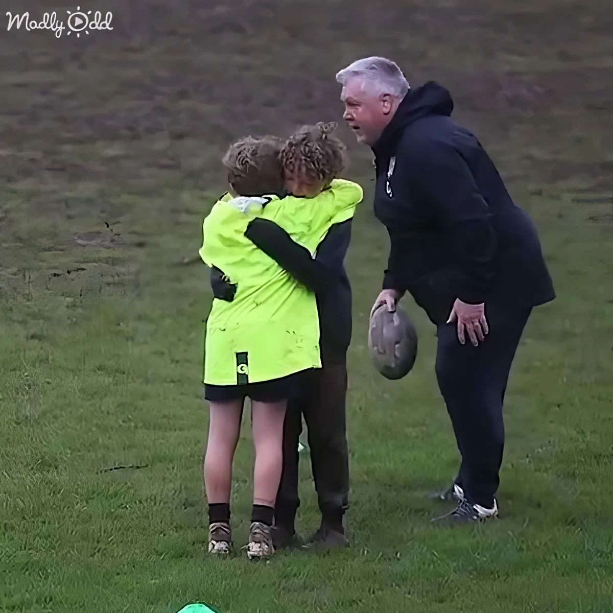 A man in black holds a rugby ball while two children in green jerseys embrace on a grassy field.
