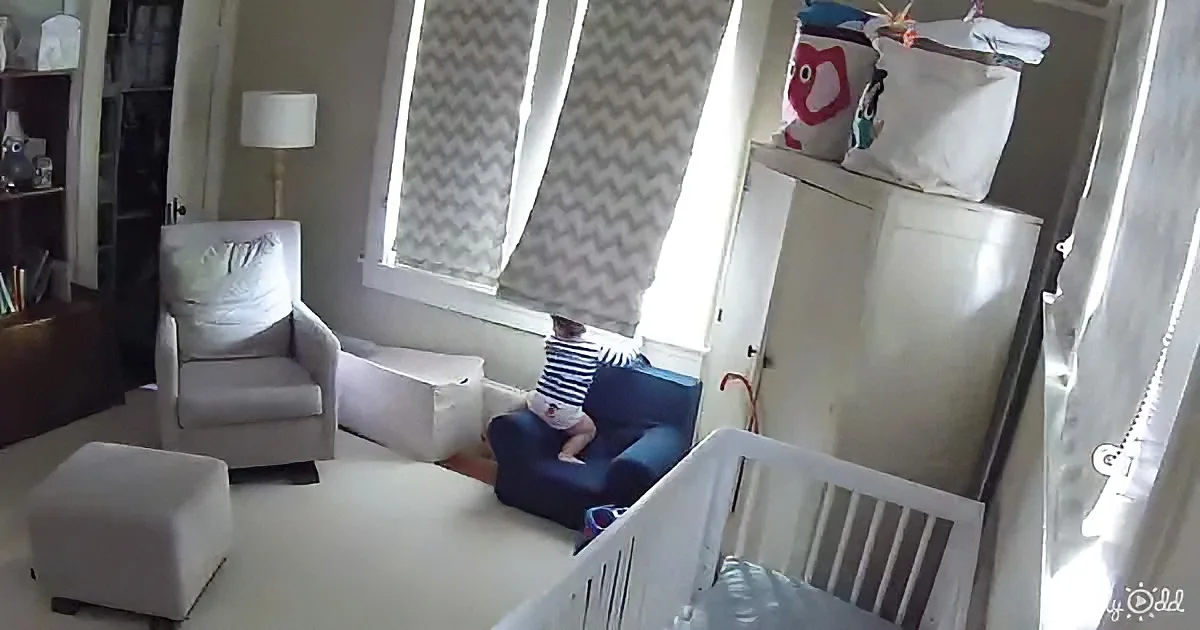 A baby in a striped shirt sits on a blue chair near a window with chevron-patterned curtains.