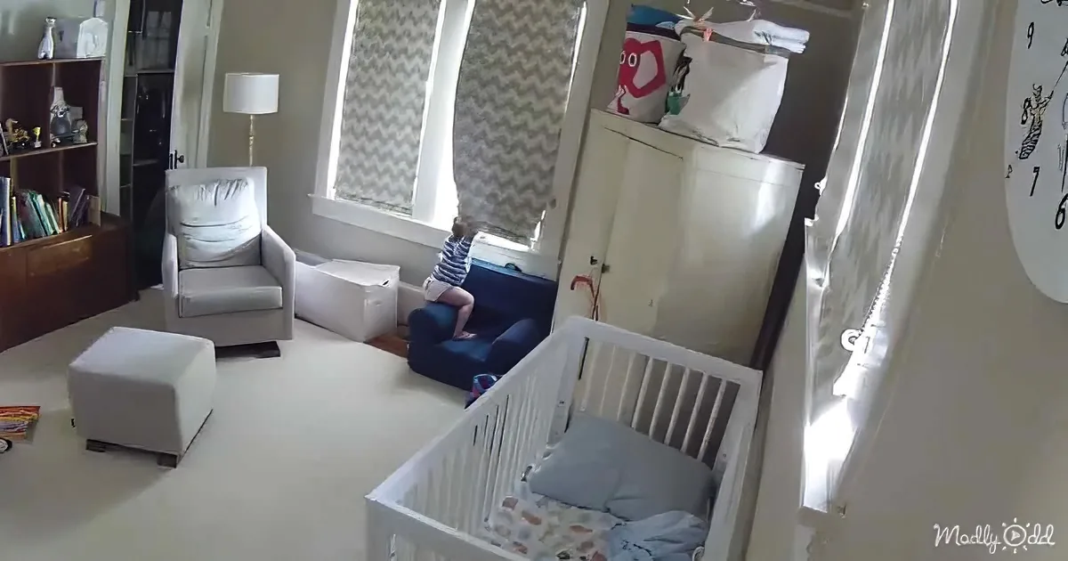 A child sits on a blue chair in a room with a crib, white furniture, and a window with chevron curtains.