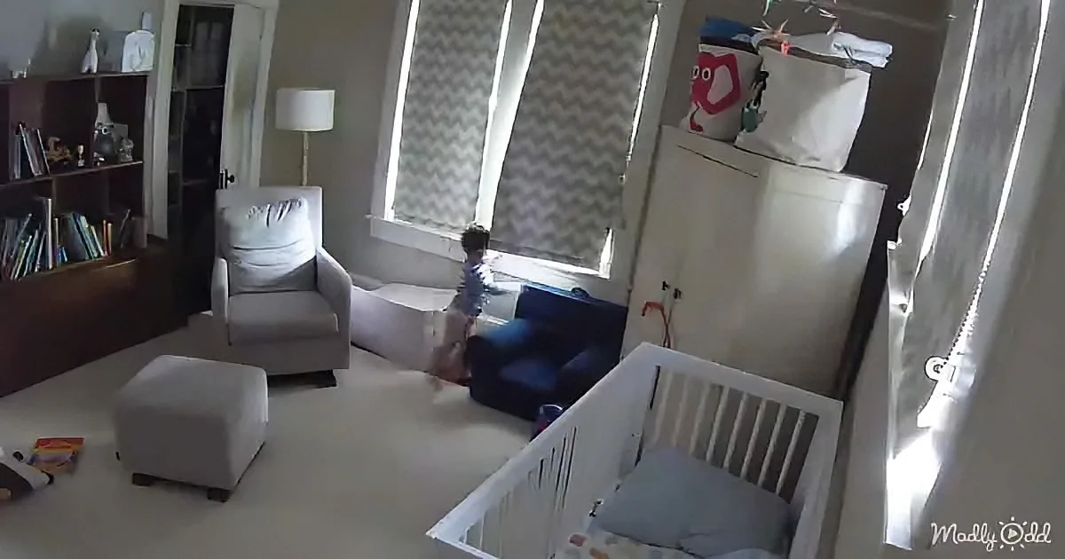 A child stands near a blue chair in a room with a crib, bookshelf, and large window.