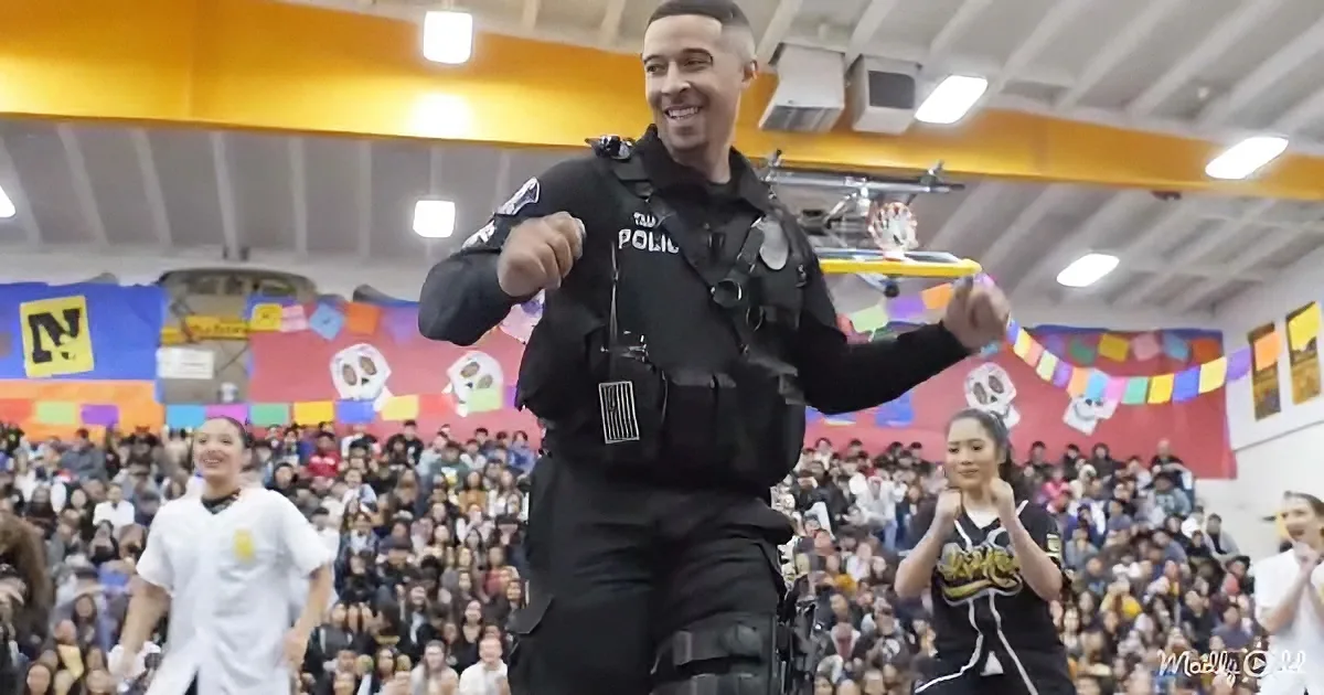 A police officer in uniform dances energetically in front of a cheering crowd in a gymnasium.