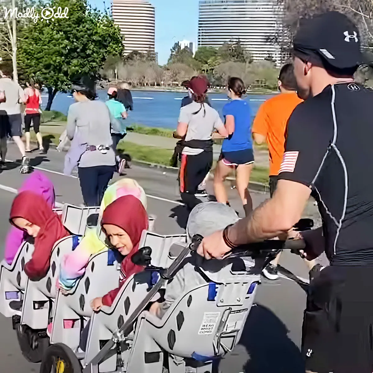 A man pushes a stroller with two children in it, walking along a path near a lake with other people in the background.