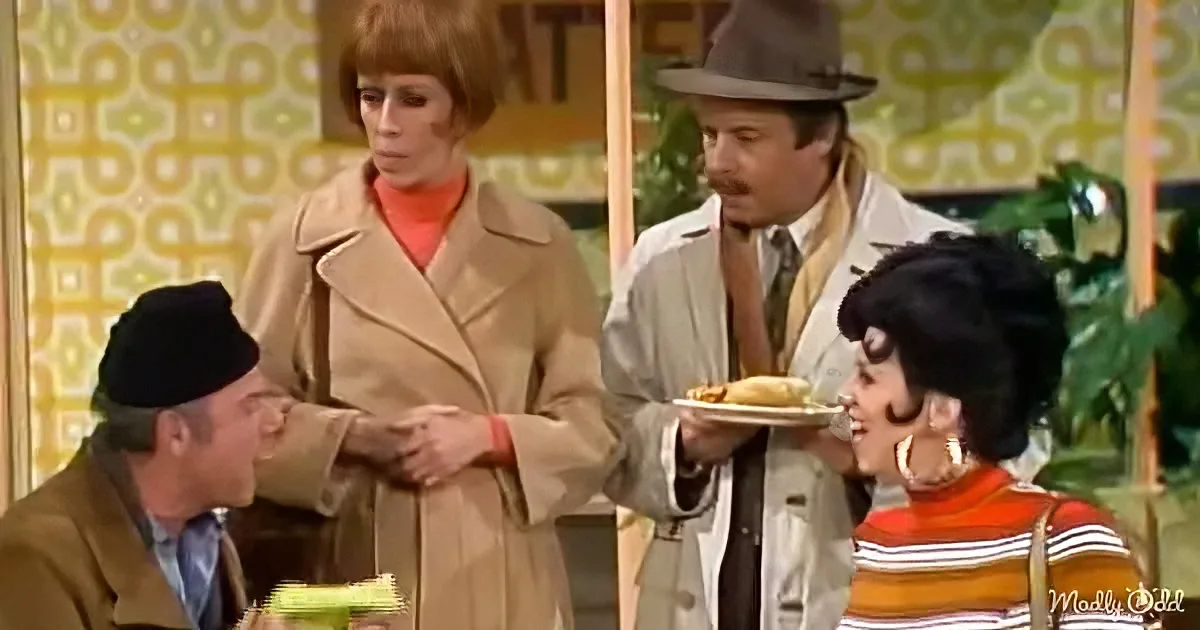 A man in a trench coat offers a plate of food to a woman with a black beehive hairstyle.