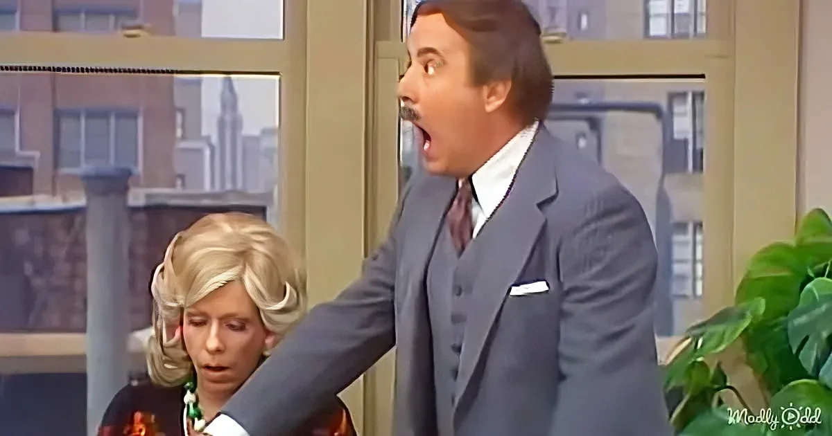 A man in a suit is surprised, mouth open, with a woman in a floral dress behind him.