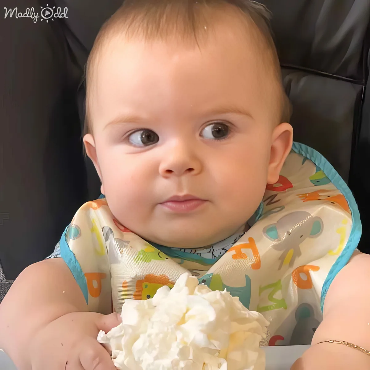 A baby wearing a colorful bib holds a large dollop of whipped cream, looking surprised.