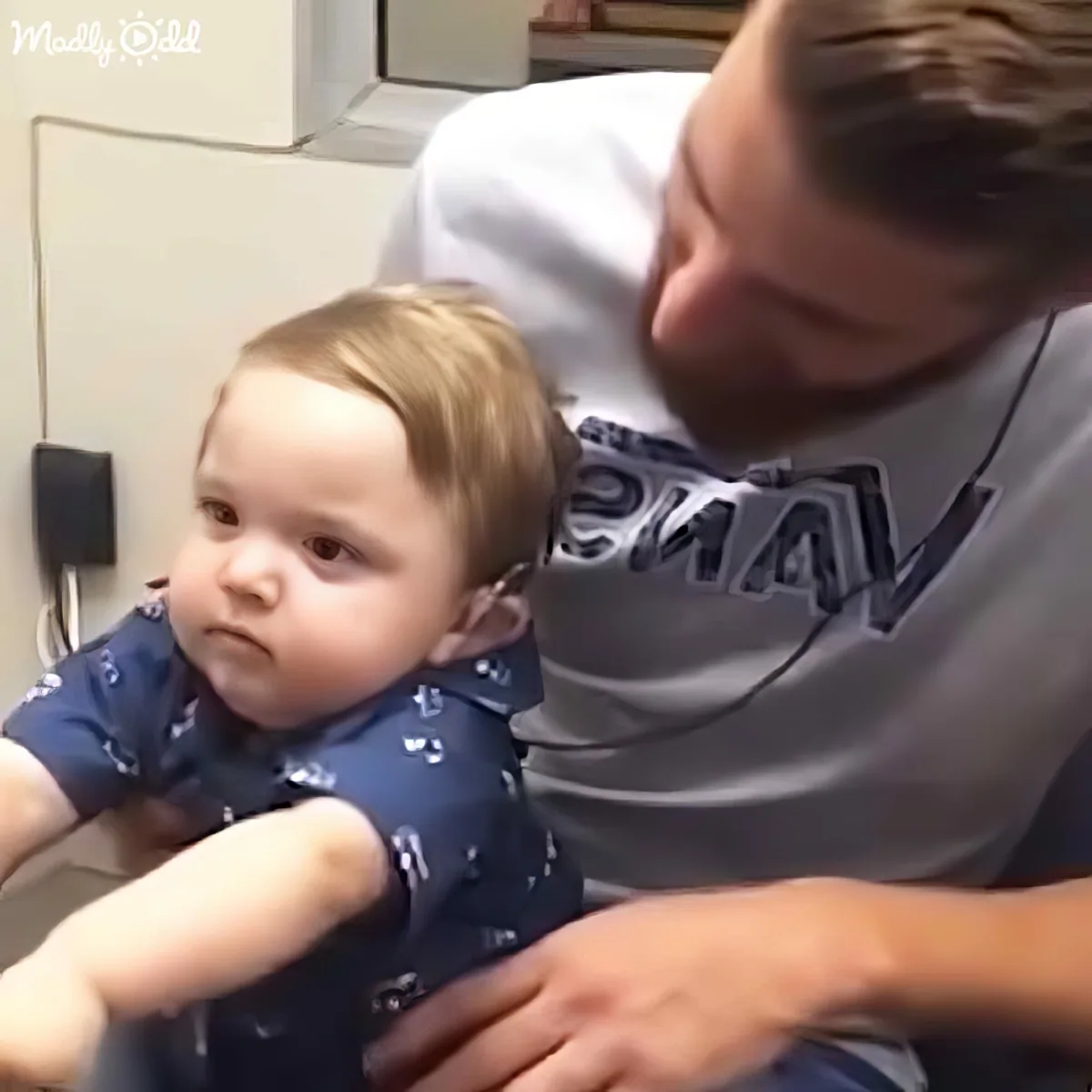 A baby in a blue outfit is being held by a person in a gray shirt in a clinical setting.