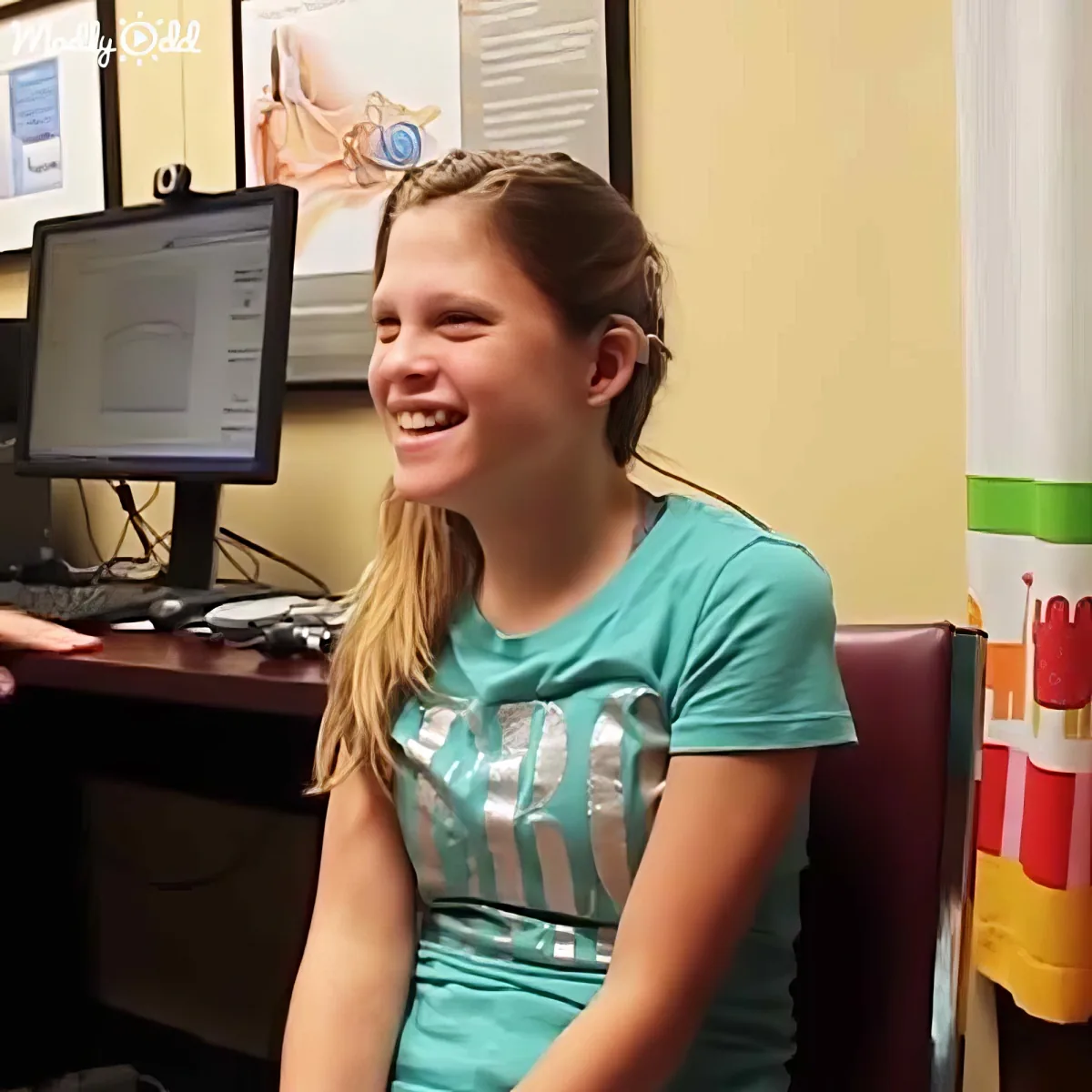 A girl in a teal shirt smiles widely, sitting in front of a computer with a colorful background.