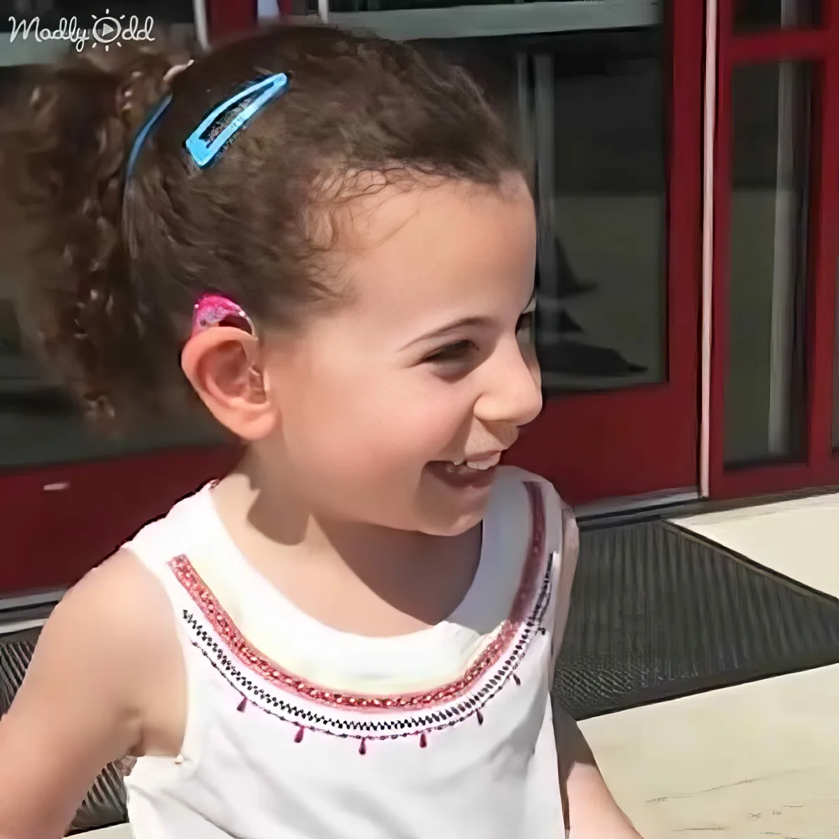 A smiling young girl with curly hair in a ponytail, wearing a white top with red trim, sits outdoors near a red building.