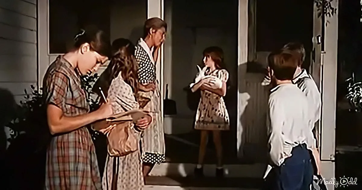 A woman in a plaid dress writes on a clipboard while standing on a porch with other people.