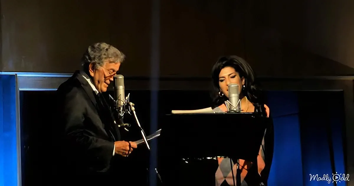 A man in a suit holds a microphone, while a woman sings at a piano with a microphone.