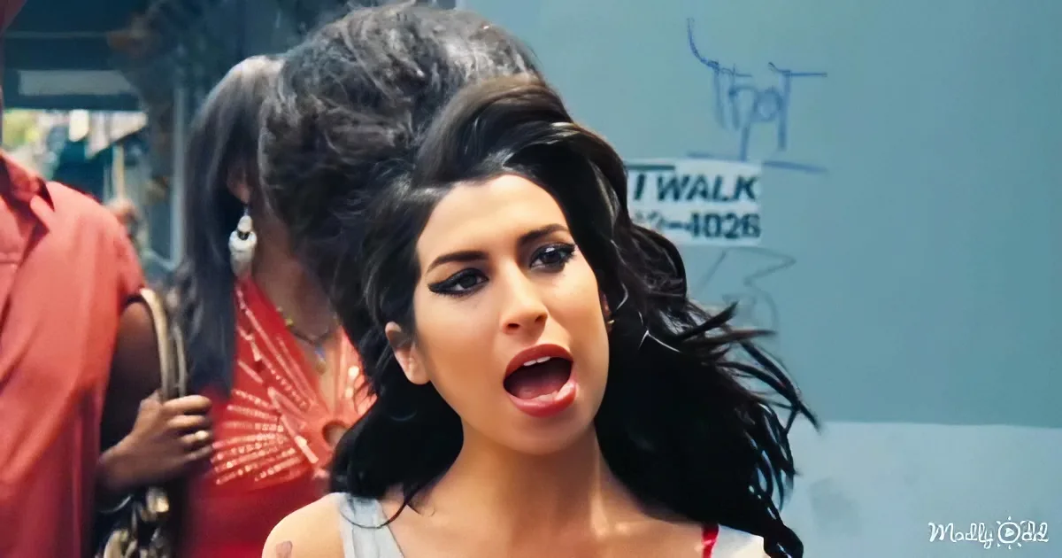 A woman with long, dark, curly hair and makeup is singing passionately, surrounded by people in a lively setting.