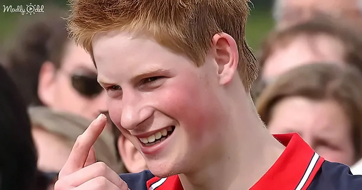 A young man with red hair smiles widely, pointing at his nose, with a blurred crowd in the background.