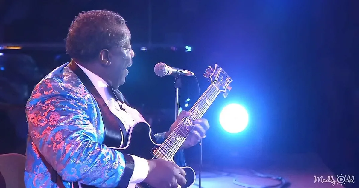 A man in a blue patterned suit plays a bass guitar on stage under blue lighting.