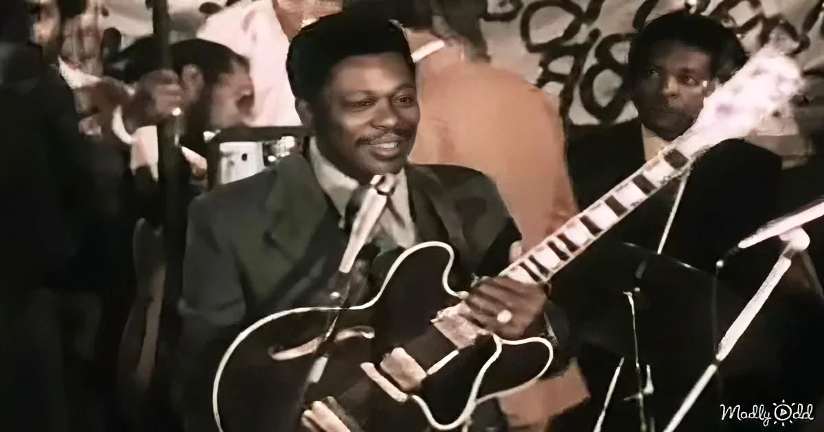 A man in a suit plays a black electric guitar in front of a crowd.
