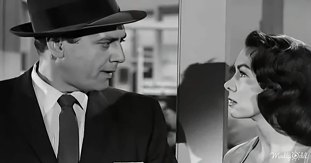 A man in a suit and hat is speaking to a woman with dark hair, both appearing serious.