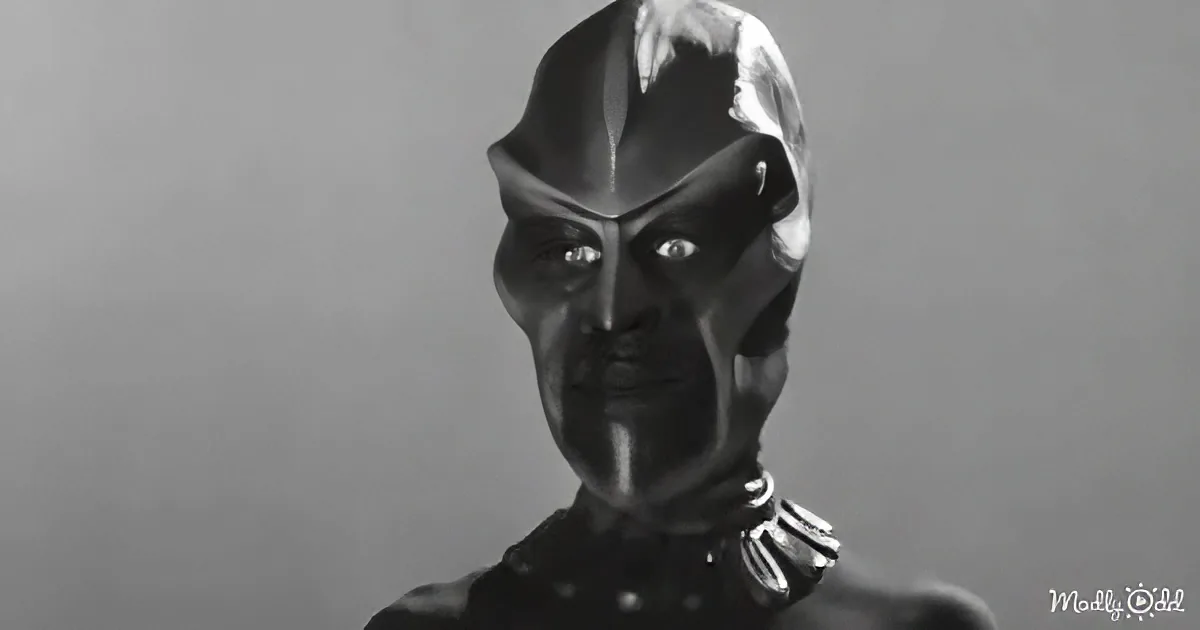 A dark, hooded figure with glowing eyes and a mask-like face stands against a gray background.