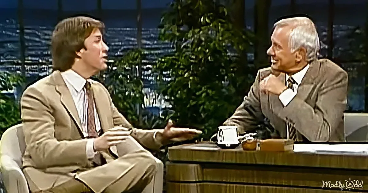 A man in a suit gestures while seated, engaged in conversation with another man in a suit, both on a talk show set.