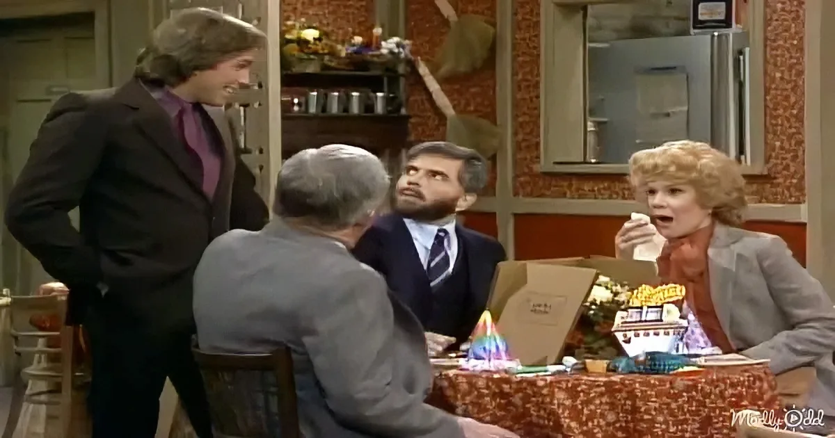 A man in a suit stands beside a table where another man looks up, while a woman reacts with surprise.
