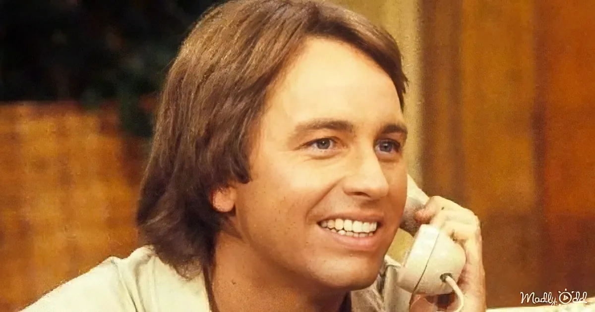 A smiling man with long hair holds a white telephone receiver, wearing a light-colored shirt.